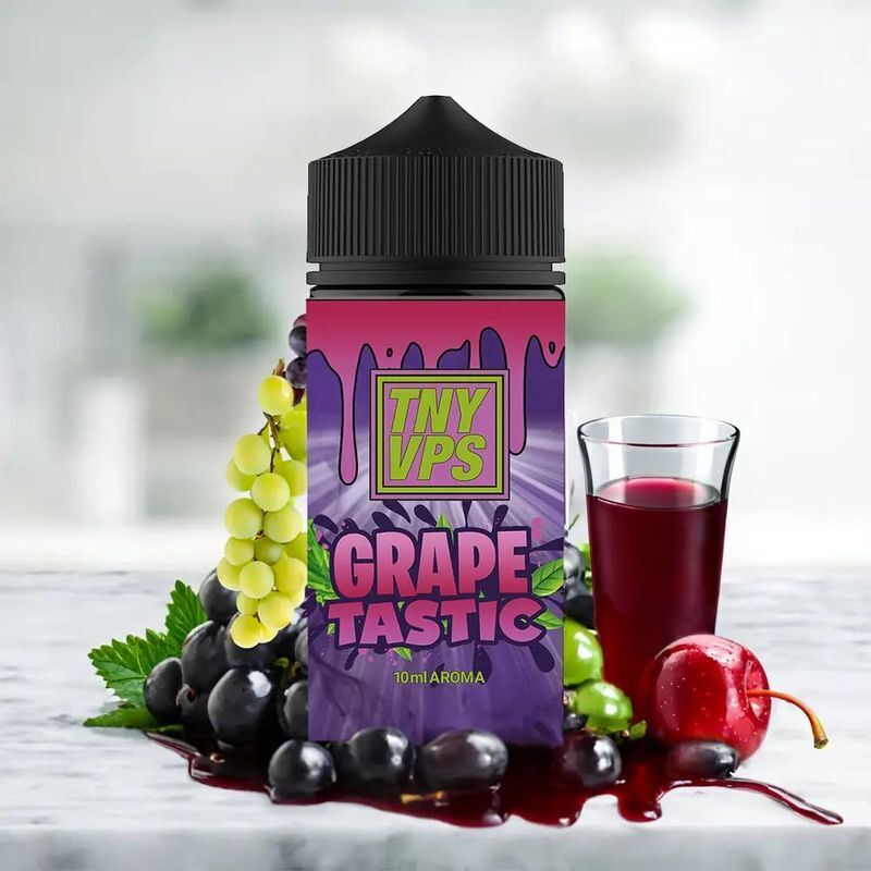 Aroma Grapetastic von Tony Vapes Flasche des Aromas Tony Vapes Grapetastic umgeben von Trauben, Kirsche und Glas mit roter Flüssigkeit auf einer Oberfläche.