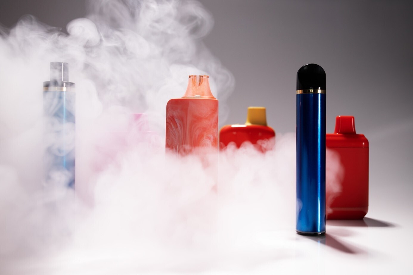 Sammlung an blauen und roten E-Zigaretten umhüllt von einer Dampfwolke vor einfarbigem, beigen Hintergrund.