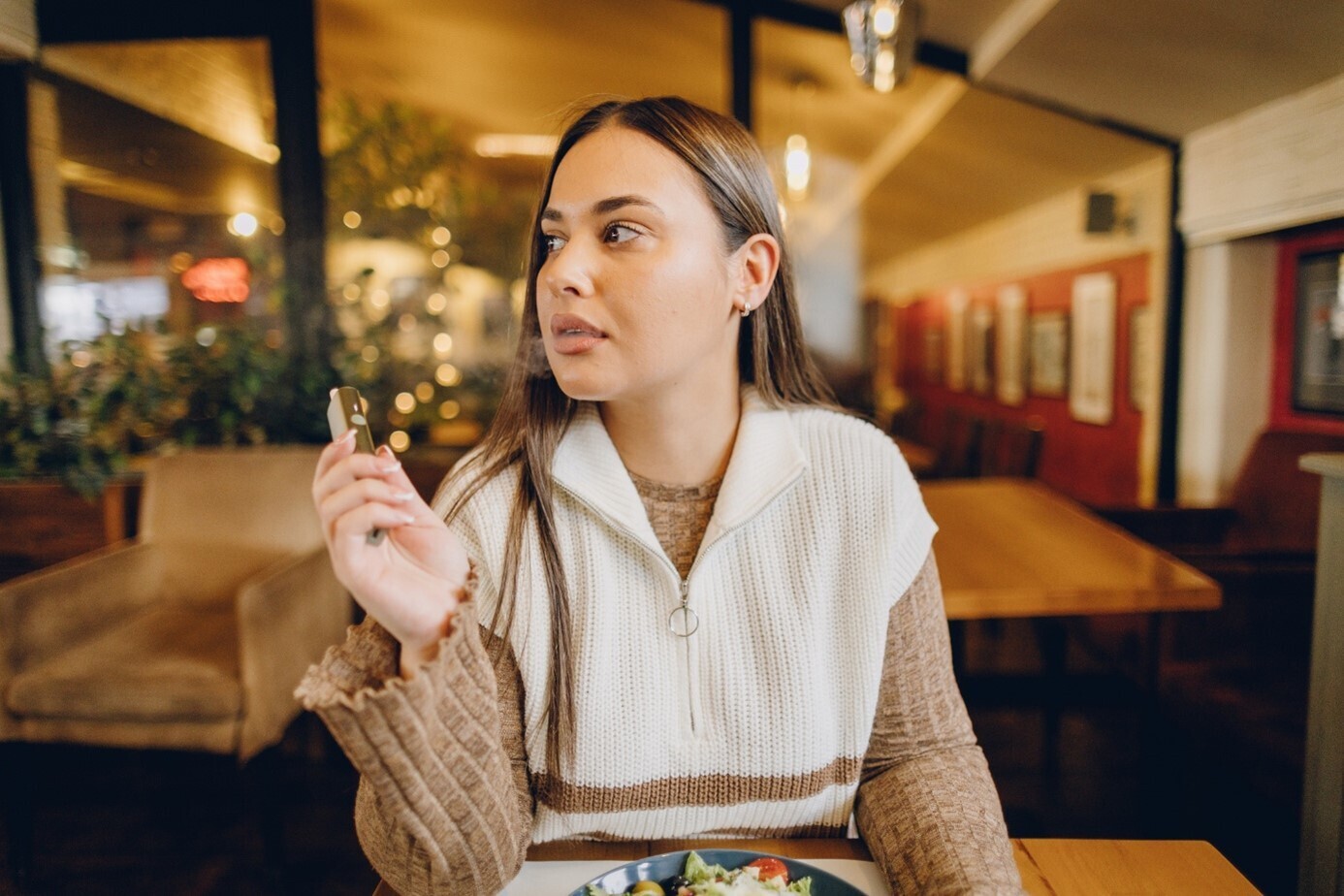 Junge Frau sitzt an einem Holz-Tisch im Coffee-Shop mit einem Salat, hält eine minimalistische E-Zigarette.