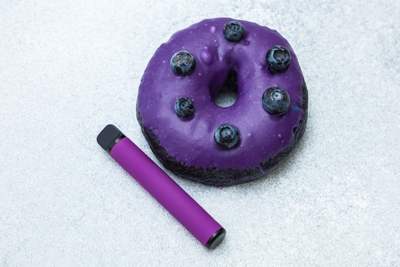 Ein Donut mit lilaner Glasur und Blaubeeren vor silbernem Hintergrund mit einem passenden, lila Vape daneben.