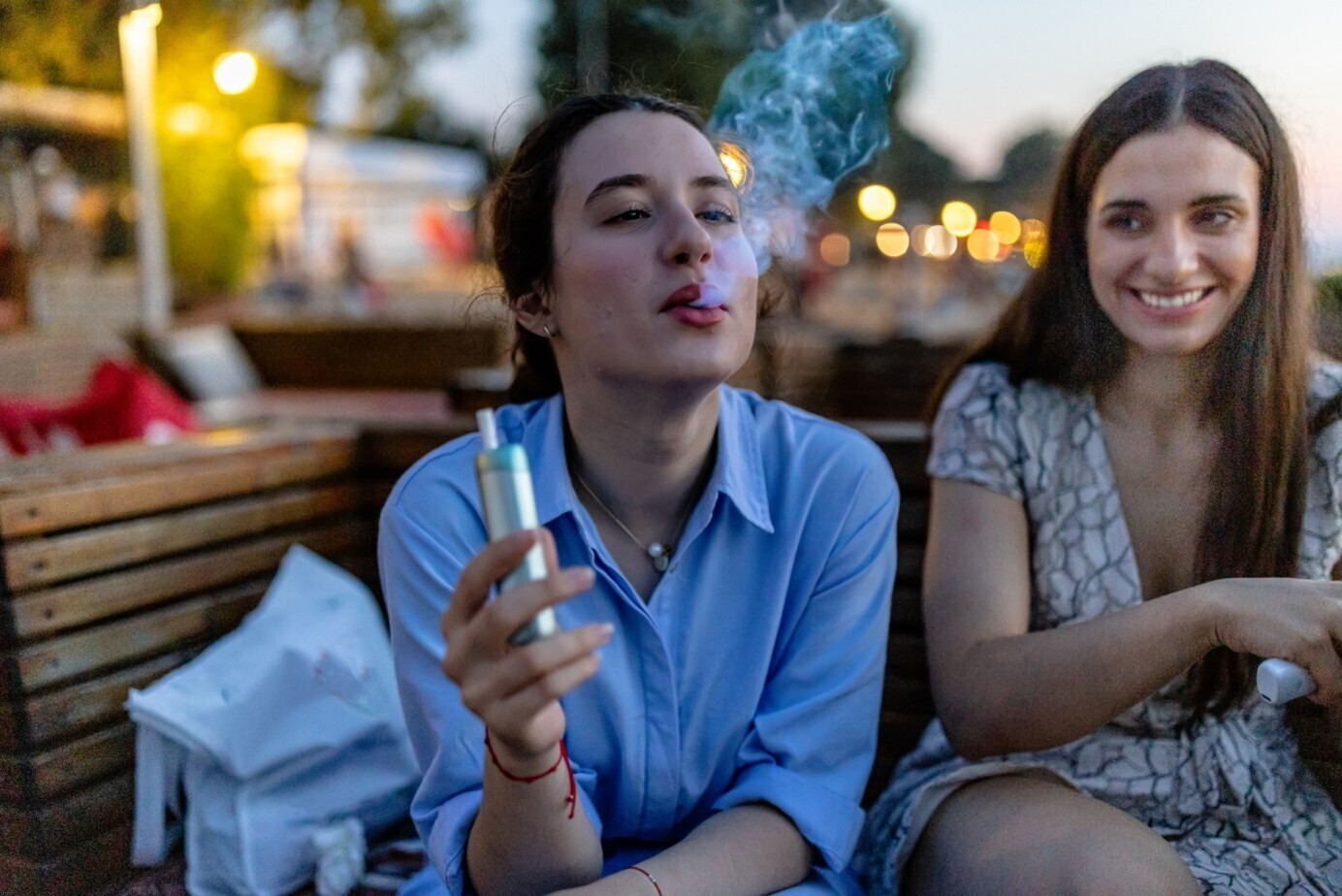 Zwei Frauen sitzen auf einer Holzbank im Freien mit ambienten Lichtern im Hintergrund, während die linke Dampft.