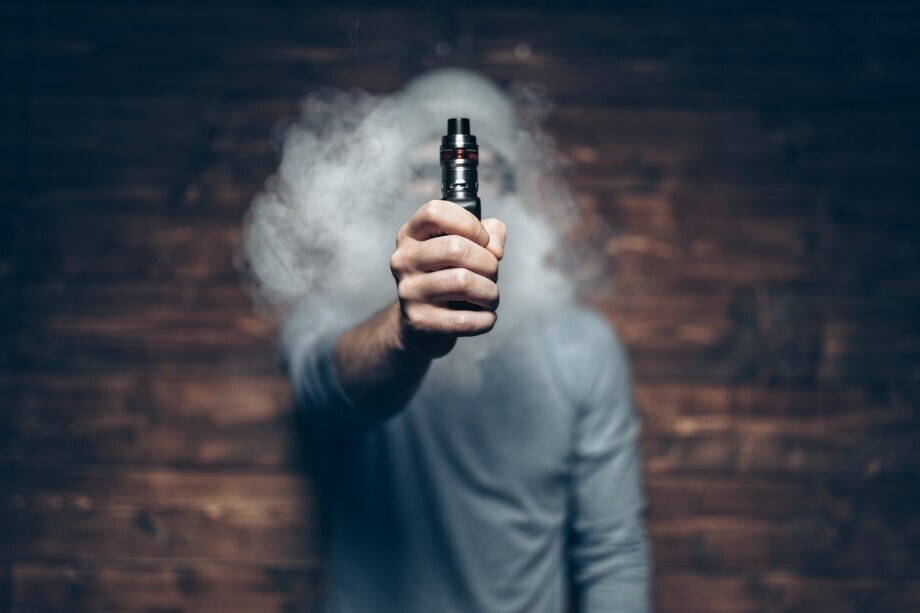 Mann, dessen Gesicht durch Dampf verdeckt ist, streckt Hand, die Vape hält, aus